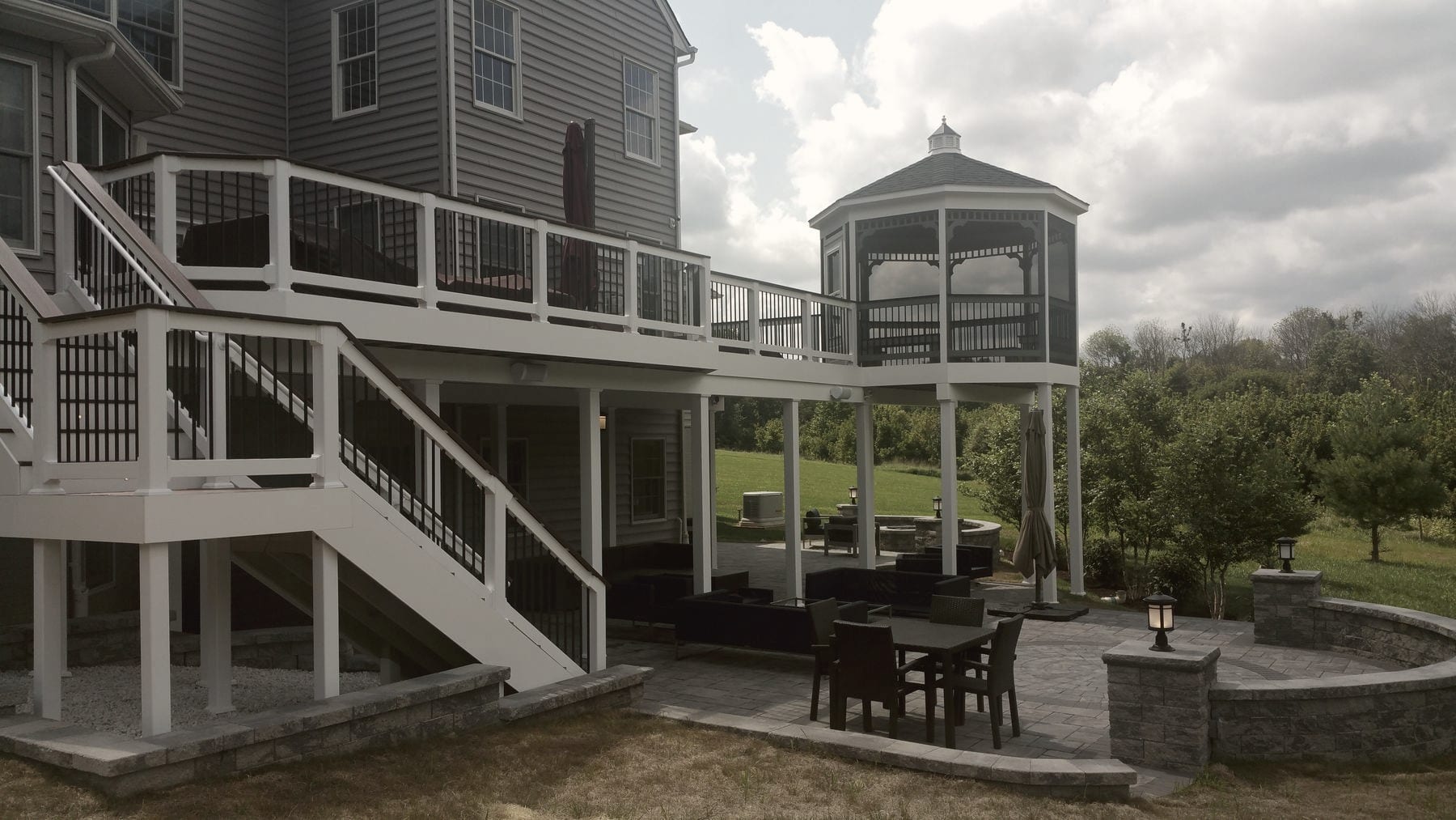 An elevated white deck with a screened gazebo, stairs down to a stone paver patio with a fire pit and curved seat walls, built by Maryland Deck Builders